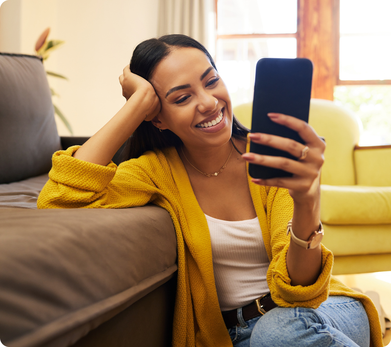 Woman in a yellow cardigan smiling at her smartphone while sitting on the floor