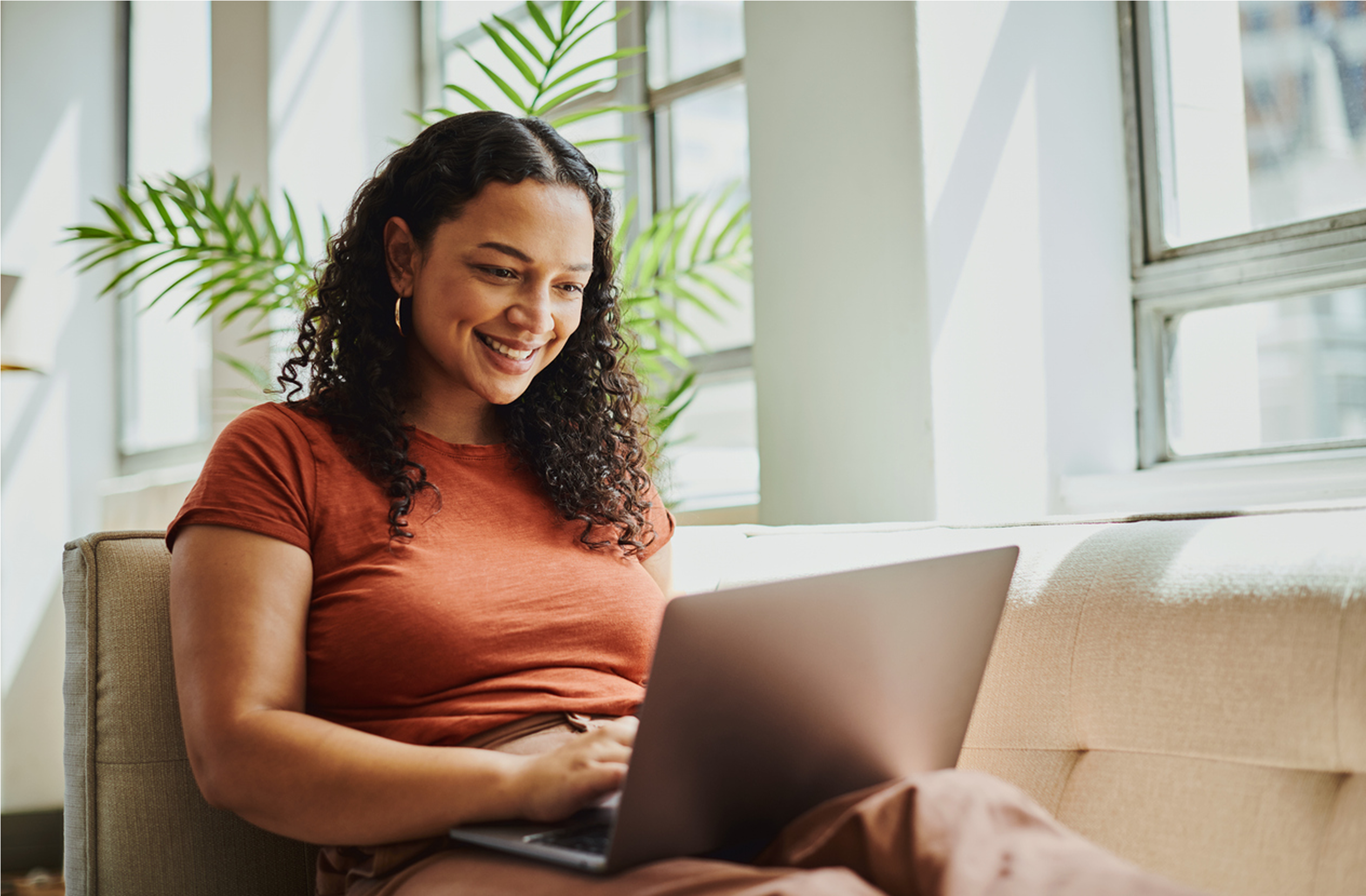Woman sitting on a sofa by large windows, smiling while working on a laptop