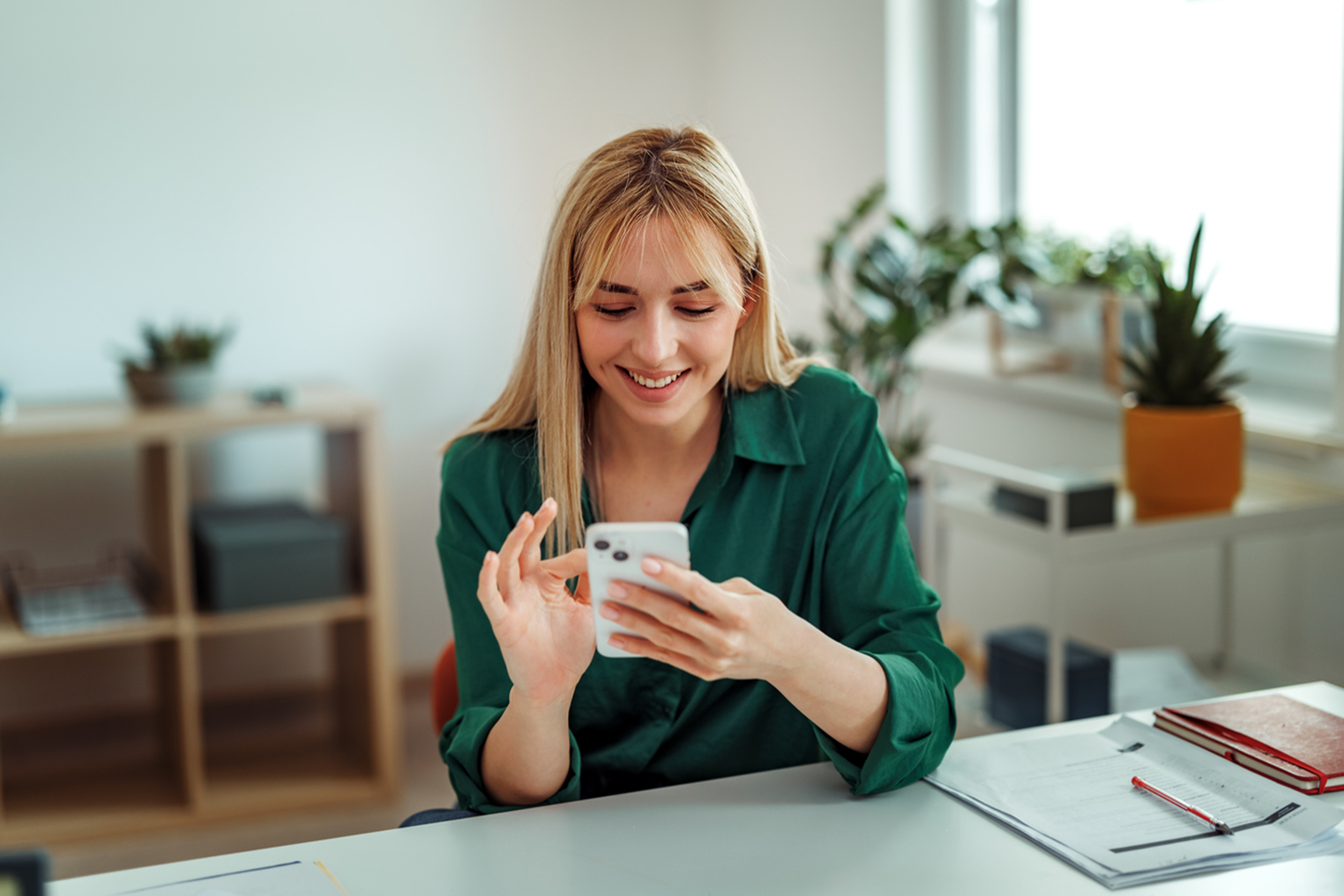 Blonde woman in a green blouse smiling while using a smartphone at her desk in a bright office